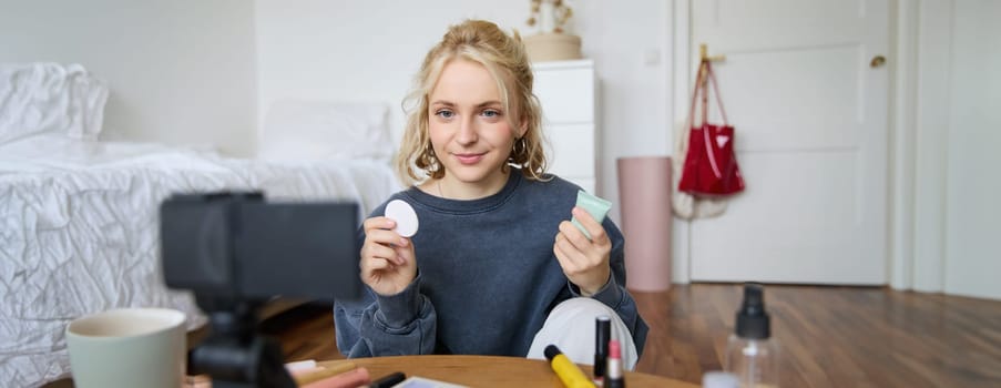 Portrait of young beauty blogger, woman gives advices to audience, how to apply makeup, sits in front of digital camera, holds foundation cream and cotton pad.