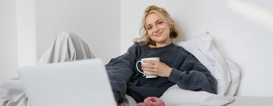 Portrait of happy woman enjoying day-off at home, lying in bed, watching tv show on laptop, looking at screen and smiling, drinking tea and eating in bedroom.