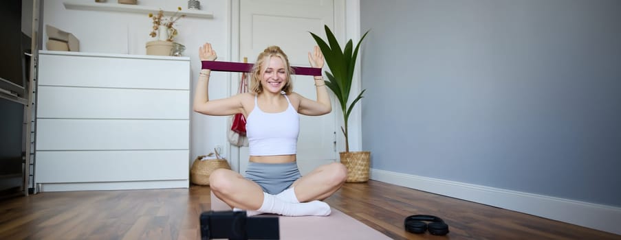 Portrait of young blond woman, social media vlogger using digital camera during workout, shooting video about exercises and fitness, stretching with rubber resistance band.