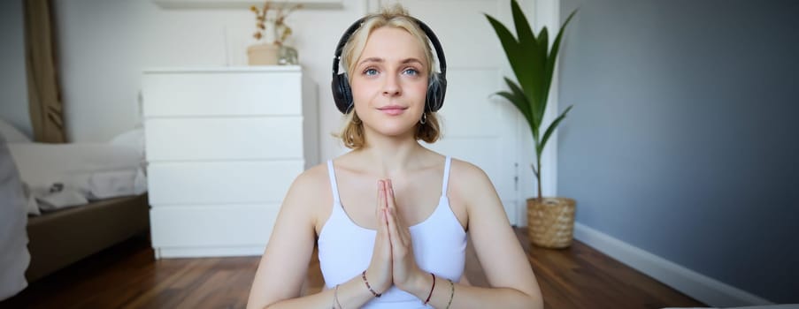 Image of young woman listens to yoga podcast for beginners, holds hands in namaste sign, wears headphones, meditates.