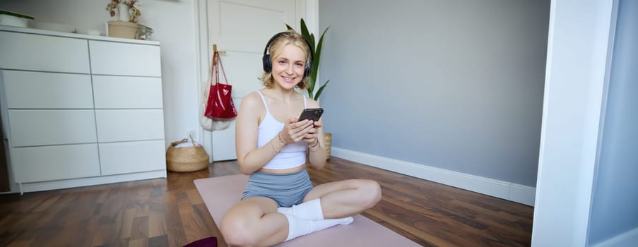 Portrait of young sporty woman in wireless headphones, sitting with smartphone on rubber mat, using workout training app, fitness application on her phone, listening in earphones.