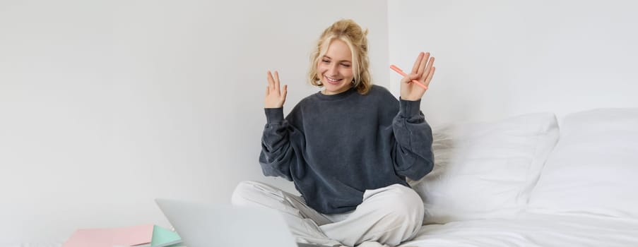 Portrait of young happy woman, connects to video chat, using laptop, waving hand at camera, saying hello to someone online, sitting on bed, studying, e-learning or working from home.