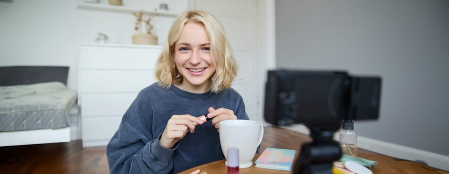 Portrait of beautiful, smiling blond woman, girl recording video of her makeup tutorial for social media, vlogger sitting on floor in her room, using stabiliser to create content, reviewing mascara.