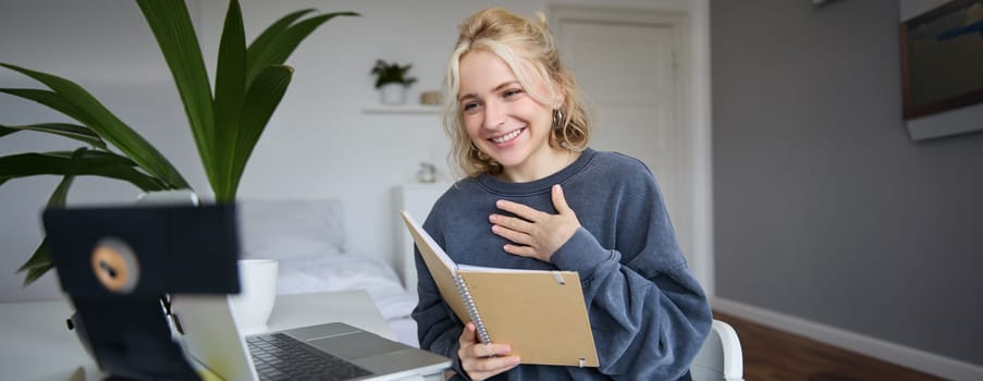 Portrait of young smiling blond woman, working from home, online chatting, using digital video camera, recording vlog, holding notebook, reading notes, explaining something.