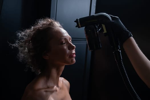 A master applies tinting paint to the face of a Caucasian woman. Instant tanning procedure.