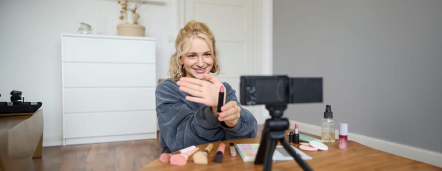 Portrait of young blond woman, teenage girl records video for her social media account, shows makeup on camera, recommends lipstick to online followers, creates content in her room.