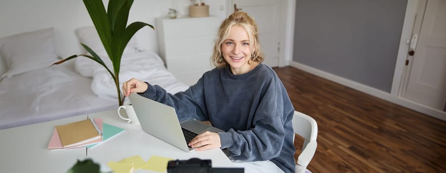 Portrait of stylish, cute young blond woman, sitting with laptop in a room, recording video on digital camera, vlogging, making lifestyle content for social media.