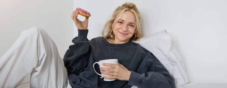 Lifestyle and people concept. Happy blond woman lying in bed with food, drinking tea and eating doughnut, resting at home, spending time alone, watching tv show.