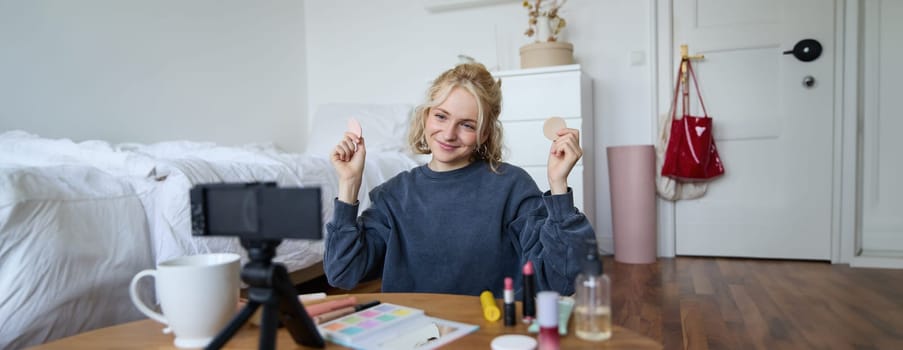 Image of young woman, vlogger records video on digital camera, shows beauty products, recommending makeup for audience on social media, sits on front in a room.