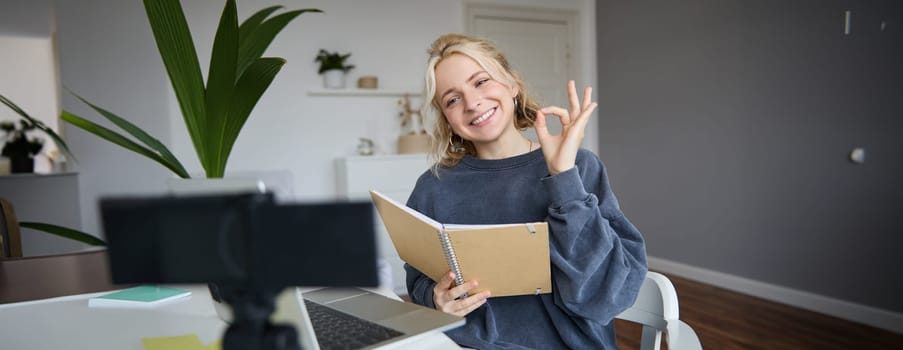 Portrait of cute, stylish young woman, shows okay sign, holds notebook, reads aloud her notes, records video vlog, looks at digital camera and makes ok gesture.