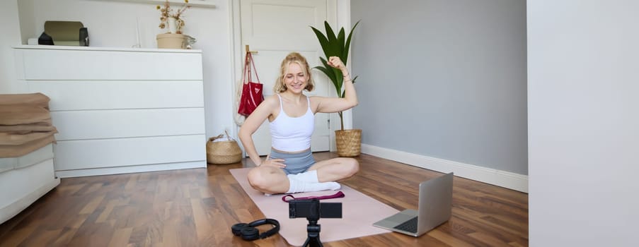 Portrait of young female athlete, fitness trainer recording vlog, training session on digital camera, sitting in a room on rubber yoga mat, showing exercises.