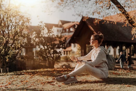 Young fashionable teenage girl with smartphone in park in autumn sitting at smiling. Trendy young woman in fall in park texting. Retouched, vibrant colors. Beautiful blonde teenage girl wearing casual modern autumn outfit sitting in park in autumn. Retouched, vibrant colors, brownish tones.