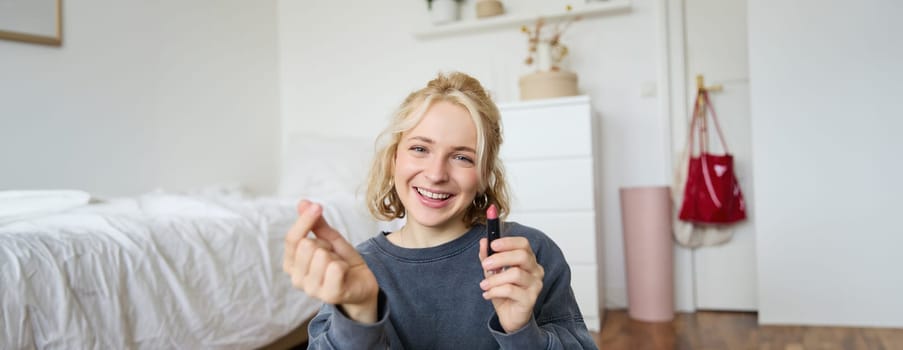 Portrait of young happy woman, content media creator recording a vlog about makeup in her room, using digital camera and stabiliser to show beauty products, holding lipstick.