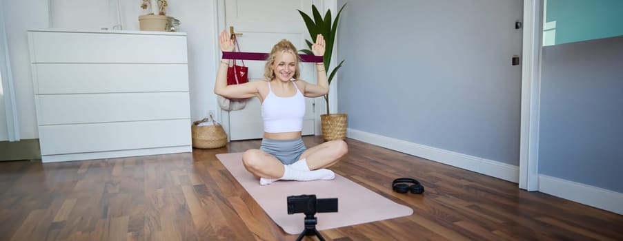 Portrait of young athletic woman recording home workout video, shooting content for sport fitness vlog, using resistance band and digital camera.