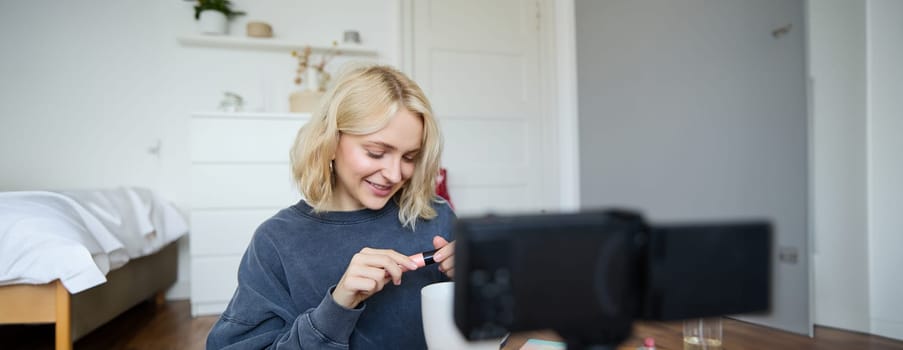 Close up portrait of smiling young woman with video camera in her room, showing mascara, reviewing beauty products for social media account. Beauty blogger creates vlog about makeup.