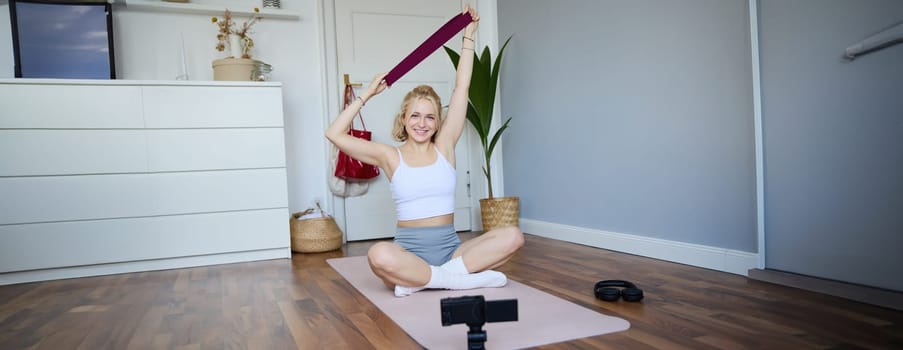 Image of smiling, athletic fitness instructor, female social media influencer, recording video about workout, showing how to use resistance bands on digital camera, sitting on rubber yoga mat at home.