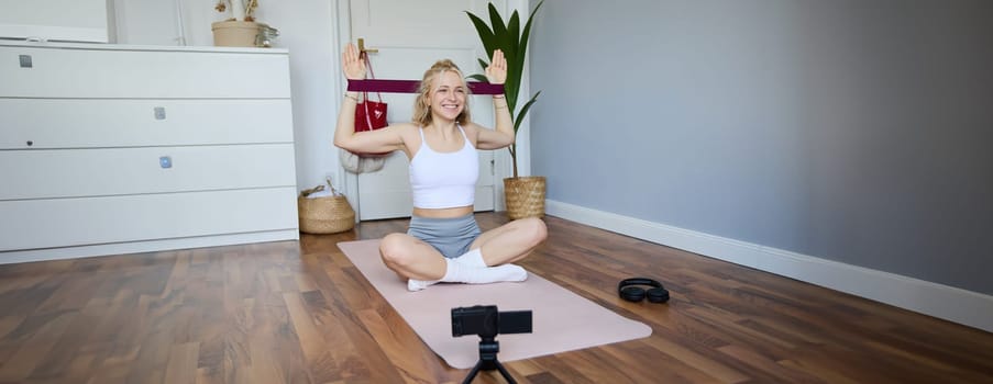 Portrait of young smiling woman, fitness instructor recording video about workout, showing how to exercise at home and use rubber resistance band, sitting on yoga mat.