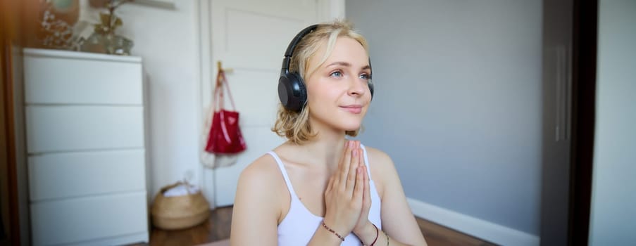 Close up portrait of young relaxed woman in headphones, holding hands together in namaste gesture, listening to meditation podcast, practice yoga.