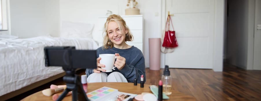 Portrait of smiling young woman, girl records video on camera, holds cup of tea, talking, doing lifestyle blog, sitting in room and creating content.