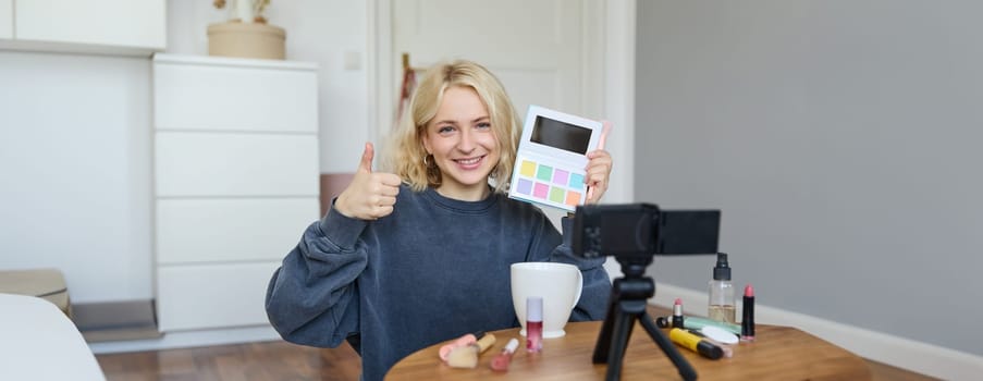 Portrait of young smiling woman, girl reviewing palette eyeshadow, shows thumbs up, recording video blog, vlogging in her room, recommending makeup product.