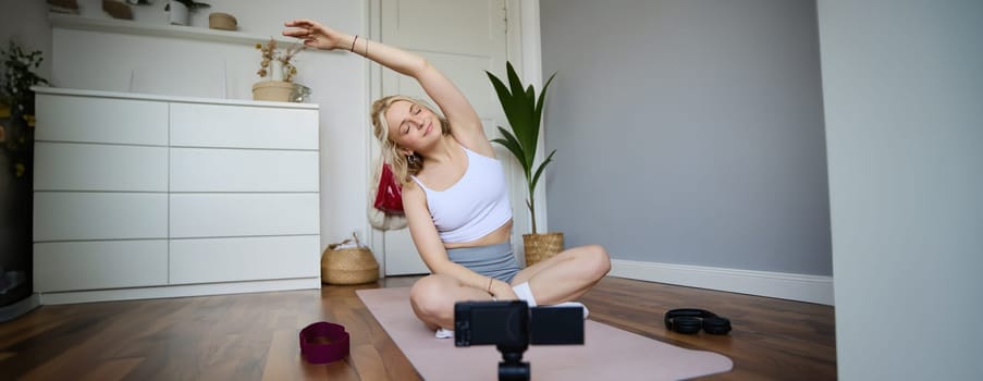 Portrait of young woman doing yoga at home, blogger recording video on digital camera while working out, personal fitness instructor showing exercises on rubber mat.
