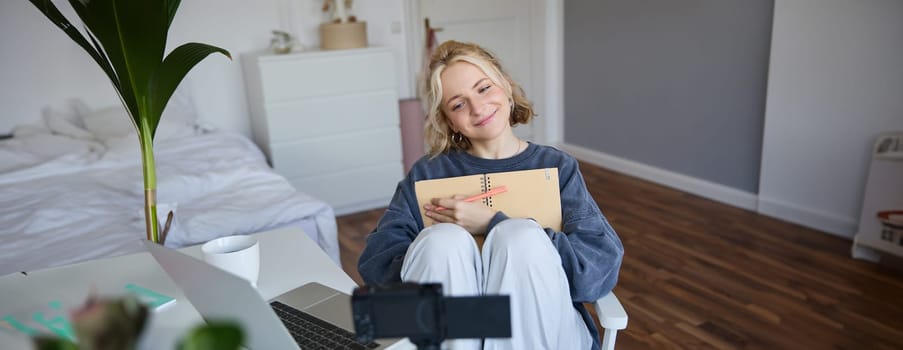 Portrait of blond smiling woman, records video on digital camera how she writes in notebook, talks to followers, doing lifestyle blog content in her room.