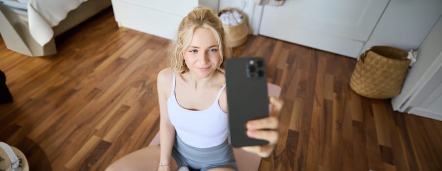 Young sporty fitness trainer, yoga instructor does live stream, takes selfies on smartphone while sitting on rubber mat at home, recording her workout training session.