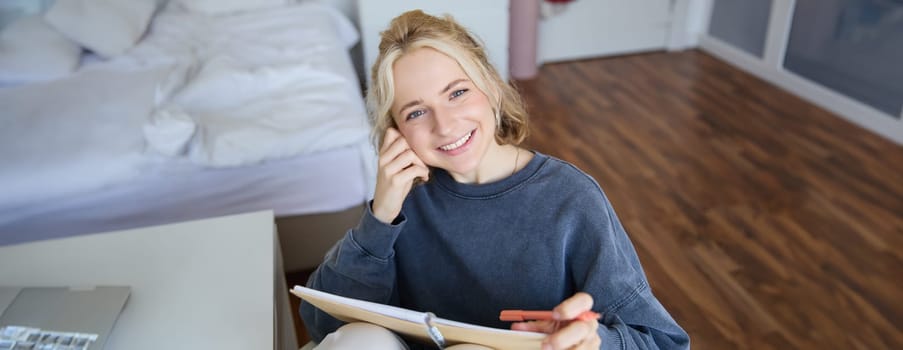 Portrait of smiling, beautiful young candid woman, sitting in bedroom with pen and notebook, writing in journal, making notes, looking happy at camera.