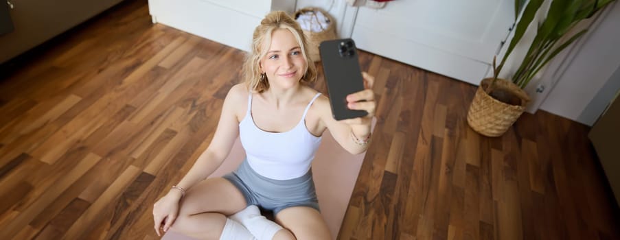 Portrait of stylish, young beautiful woman takes selfie while working out at home, using smartphone to make photos on yoga mat, posing in fitness clothes.