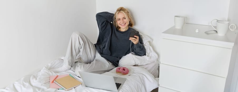 Portrait of young woman, student studying in her bed, relaxing while preparing homework, eating doughnut, using laptop in bedroom and drinking tea.