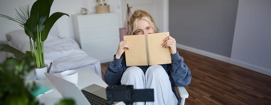 Portrait of cute, smiling young social media content creator, girl records video on digital camera and stabiliser, holds notebook, talks to audience, vlogging in her room.