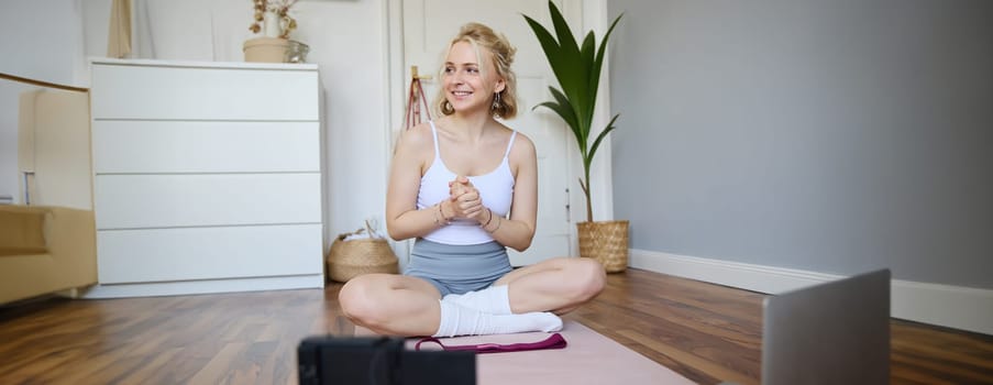 Portrait of young fitness instructor, vlogger showing exercises on camera, recording herself, sitting on mat with laptop, doing workout, explaining yoga movements to followers.