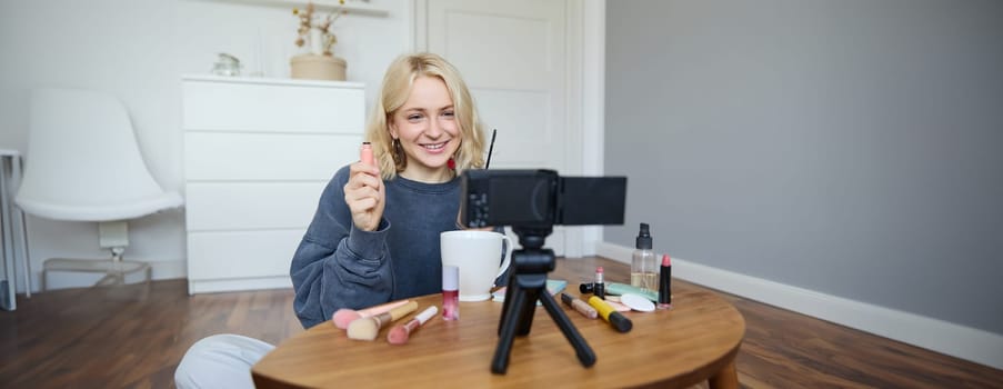 Portrait of beautiful, smiling blond woman, girl recording video of her makeup tutorial for social media, vlogger sitting on floor in her room, using stabiliser to create content, reviewing mascara.