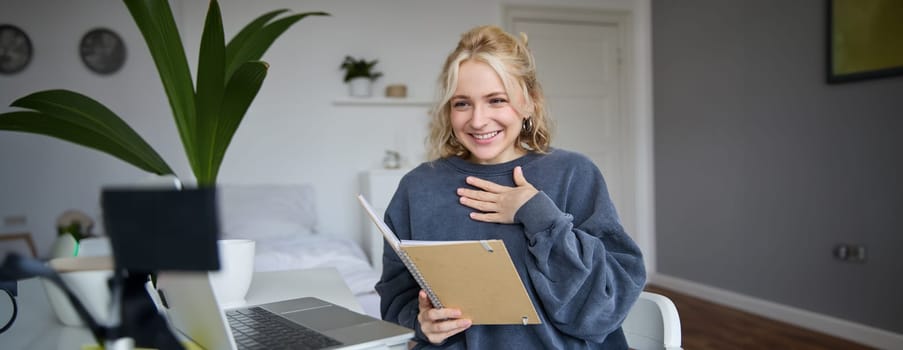 Portrait of smiling young cute woman, holding notebook, girl with laptop and planner in hands, sits in room, looks at digital camera, creates lifestyle vlog, talks to audience, reads script.