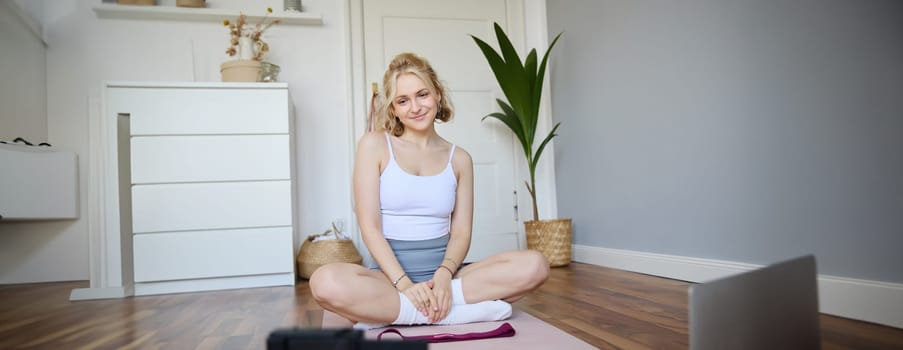 Portrait of young fitness instructor, vlogger showing exercises on camera, recording herself, sitting on mat with laptop, doing workout, explaining yoga movements to followers.