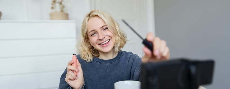 Image of cheerful, beautiful young lifestyle blogger, woman sitting on floor and recording video about makeup, holding mascara, making lifestyle content for her social media account and followers.