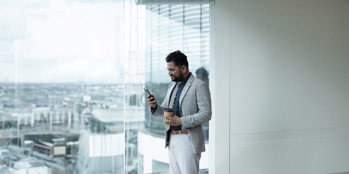 Indian businessman standing next to window using mobile phone while coffee break.