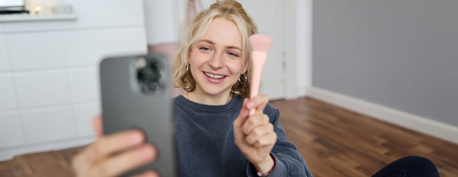 Image of stylish young woman, social media influencer, taking pictures on mobile phone, doing makeup tutorial for followers online, recording video vlog in her bedroom, showing brush.