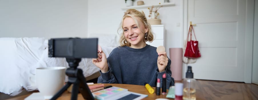 Portrait of young woman, beauty content creator, sitting in a room in front of digital camera, recording makeup tutorial vlog, showing cosmetic facial products.