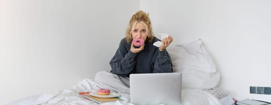 Portrait of sad woman crying, eating doughnut, wiping tears off, looking at something upsetting on laptop screen, sitting on a bed.