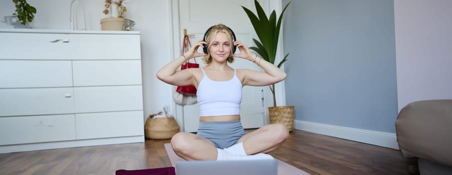 Portrait of young fitness girl, woman workout at home, listens to fitness instructor in wireless headphones, follows exercises on laptop.