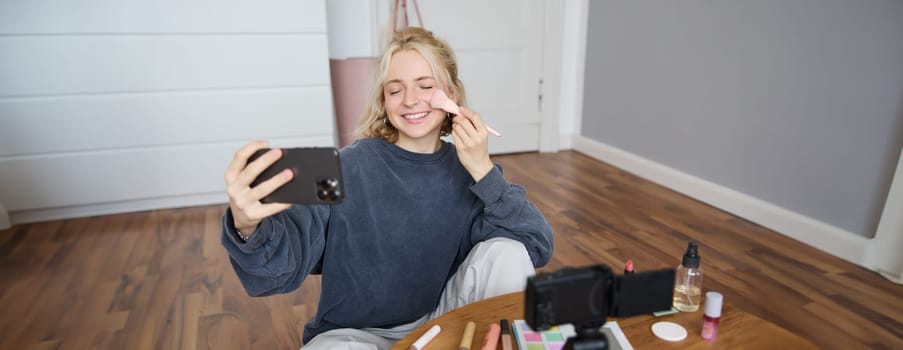 Image of young woman, vlogger taking selfie in her room, talking to her followers during online live stream, using smartphone app to chat with audience, smiling and looking happy.