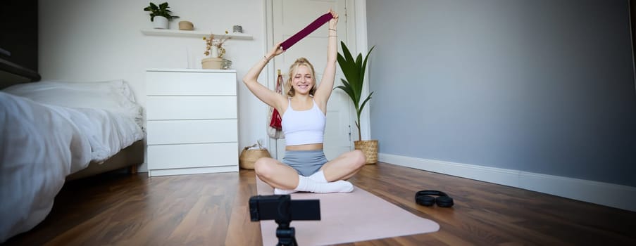 Smiling young blond woman recording video about workout, shooting on digital camera, fitness instructor shows how to use resistance band during exercises. Lifestyle and wellbeing concept
