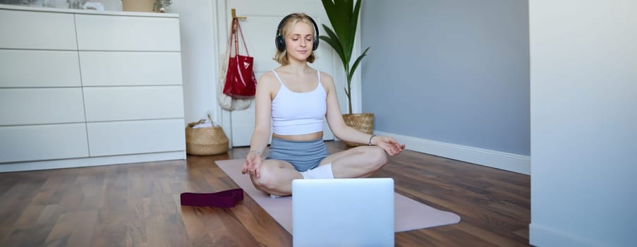 Portrait of young and relaxed woman using laptop and wireless headphones while meditating, practice yoga at home on rubber mat, sitting in asana.