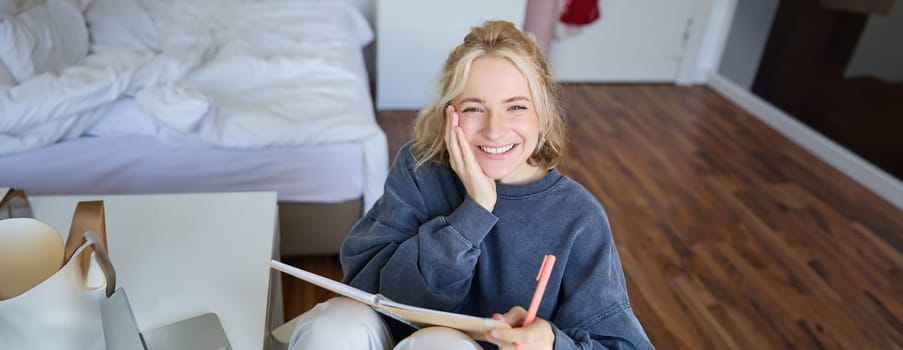 Portrait of smiling, charismatic young woman, writing down notes, making plans and putting it in planner, holding journal, sitting in bedroom and looking happy at camera.