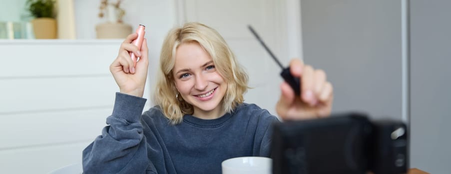 Portrait of young smiling woman in her room, recording video on camera, lifestyle vlog for social media, holding mascara, reviewing her makeup beauty products, showing how to use cosmetics.