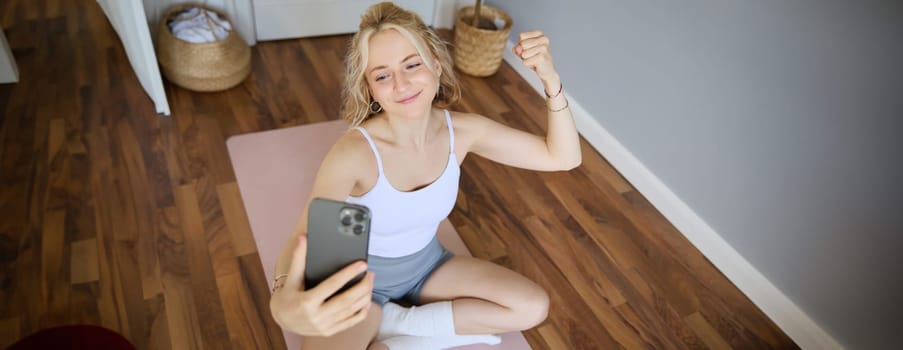 Portrait of young sporty woman taking selfie during workout training, content creator takes photos, shows muscles on camera, flexing biceps.