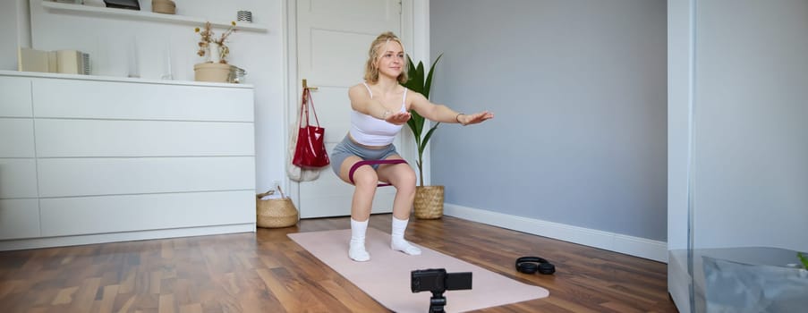 Portrait of young woman, fitness instructor making a video for sport vlog, doing squats on camera, using resistance band, working out at home.