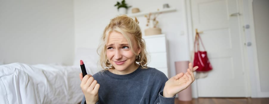 Portrait of woman, vlogger looking disappointed, showing lipstick and shrugging shoulders, recording video about makeup for social media account.