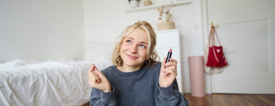 Portrait of cute, charismatic beauty blogger, woman sits in a room with lipstick in hand, talking about makeup, chatting with followers, recording online stream on social media app.